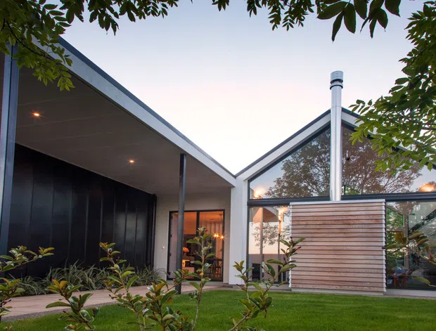 Contemporary single-storey residence viewed from a landscaped garden, featuring a dramatic asymmetrical pitched roof, dark vertical wall cladding, slim metal framing, extensive glazing, and a timber slatted privacy screen around the entry or outdoor area. Warm interior lighting is visible through the glass, with mature foliage framing the view.