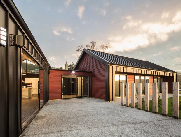 Exterior view of a contemporary single-storey building arranged around a paved courtyard. The composition features dark vertical metal cladding and extensive glazing on the left, a red timber-clad gabled wing in the centre, and a long dark pitched metal roof. A timber-and-metal post fence defines the lawn edge, with warm interior lighting visible through the windows.