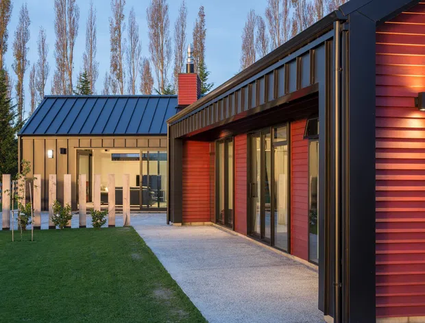Exterior view of a contemporary single-storey residence with dark standing-seam metal roofs, red horizontal cladding, extensive glazing, and a covered paved walkway opening onto a lawn. Tall trees form the background, while warm interior and exterior lighting create contrast against the cool evening setting.
