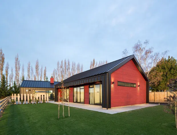 Exterior view of a contemporary single-storey house with a bold red weatherboard-clad gabled volume, dark metal roofing and trim, and large glazed openings along the side. The home sits within a landscaped lawn and fenced garden, with a connected lower wing visible to the left.