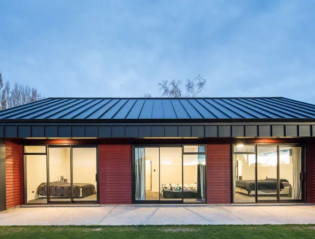 A modern single-storey residential building viewed from the exterior at dusk. The facade combines dark metal cladding and a standing-seam metal gable roof with red vertical timber or composite wall panels, large floor-to-ceiling glazed openings, and warm interior lighting. A paved terrace runs along the front, with landscaped lawn and fencing visible around the building.