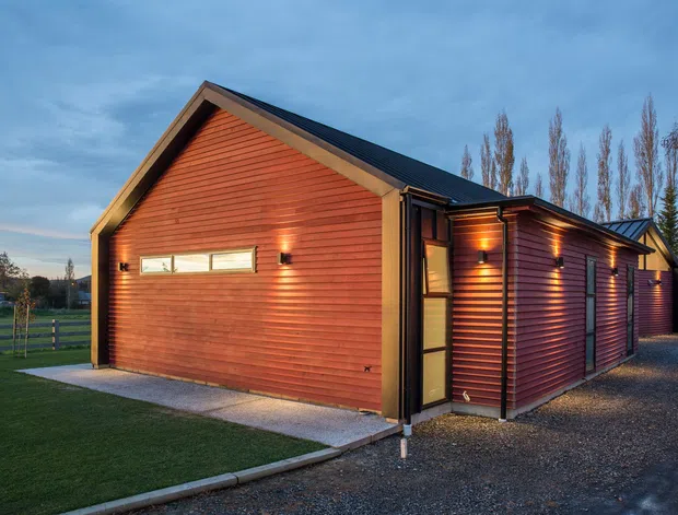 Exterior view of a contemporary single-storey building clad in warm horizontal timber weatherboards, with a prominent gabled end, long pitched roof, narrow clerestory-style window, dark trim, and warm exterior lighting. The building sits beside a gravel driveway and landscaped lawn in a rural or semi-rural setting.
