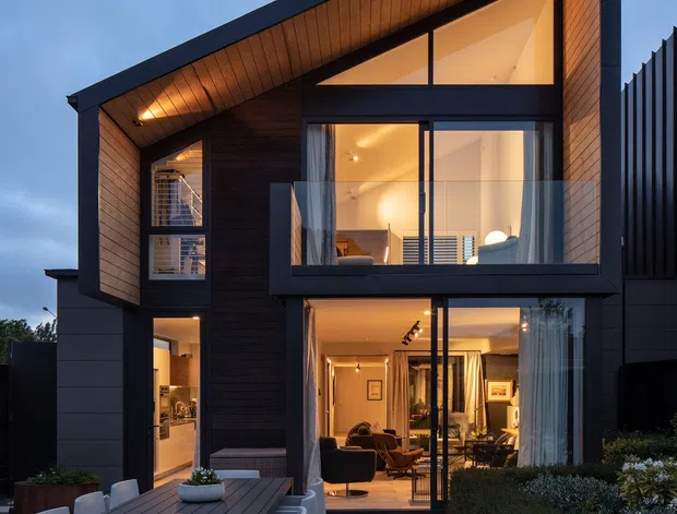Twilight exterior view of a contemporary multi-storey home with a dramatic asymmetrical pitched roof and deep timber-lined eaves. Dark cladding and black window frames contrast with warm interior lighting visible through expansive glazing. A paved outdoor dining terrace with a large table and chairs occupies the foreground, with landscaped garden beds alongside the house.