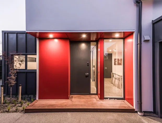 Contemporary residential entrance viewed from outside, featuring a compact timber deck, bold red recessed entry surround, matte dark front door, glazed sidelight, recessed downlights, and dark vertical cladding. The entry opens toward a bright interior hallway or living space.