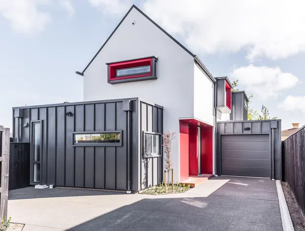 A compact contemporary multi-storey urban home with a sharply pitched gable roof, white rendered upper walls, and a dark grey vertically panelled front extension. Bold red window surrounds and a red recessed entry add contrast. The property includes a single garage, narrow paved driveway, timber boundary fencing, and a small landscaped planting strip.