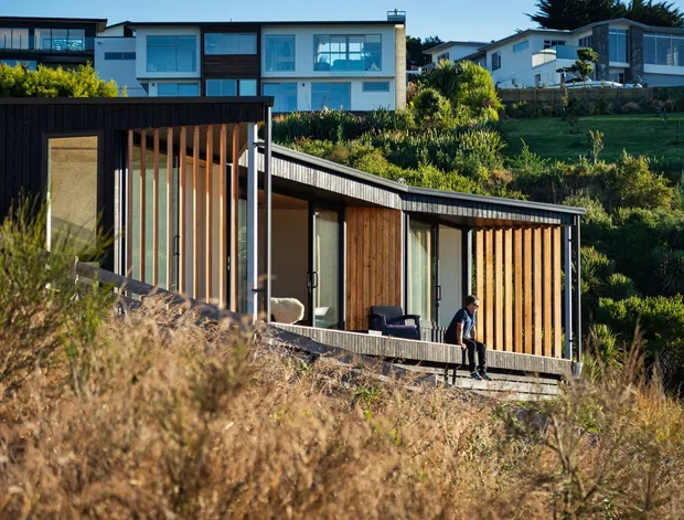 Exterior view of a compact contemporary dwelling set into a sloping, landscaped coastal-looking site. The foreground building has a long low form, dark cladding, extensive glazing, vertical timber screening and a single sloping roof, with a sheltered deck or outdoor seating area. Larger modern homes are visible upslope in the background.