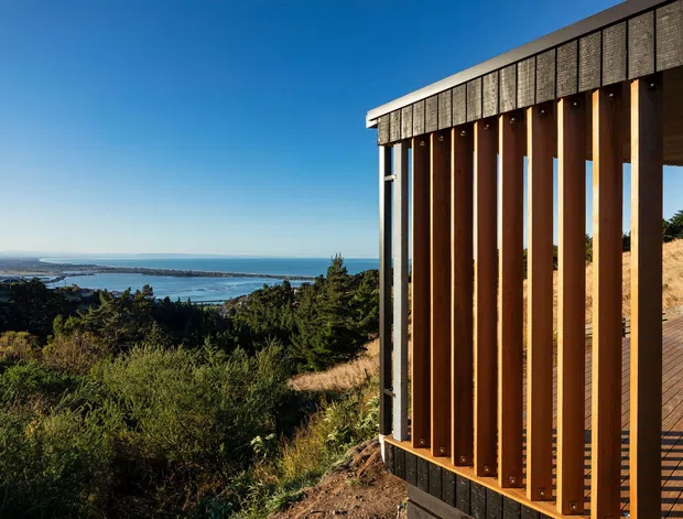 Exterior architectural view of a contemporary structure on a sloped, vegetated site overlooking a broad coastal inlet or bay. The building features dark vertical cladding or screening, exposed warm timber vertical fins, and a narrow timber deck or walkway, with expansive coastal scenery beyond.