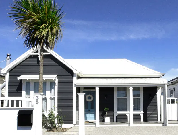 A bright street-facing exterior of a compact single-storey home with dark horizontal weatherboard cladding, crisp white trim, a front gable, and a low-pitched main roof. The entry porch has white posts and railings, with a small paved forecourt and a tall palm tree beside the facade.