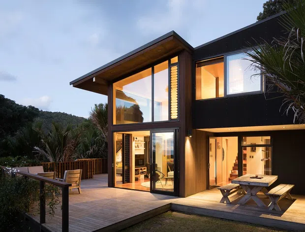 Exterior view of a contemporary two-storey house set among dense vegetation, with bold black cladding, extensive floor-to-ceiling glazing, projecting flat roof planes, and warm interior lighting. A timber deck wraps around the building, including an outdoor dining area with a picnic table and chairs, while glass balustrades and timber screening define the outdoor spaces.