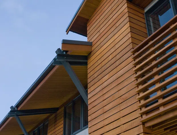 Upward-looking architectural exterior detail of a contemporary multi-level building. The facade combines warm horizontal timber cladding, exposed grey structural beams, deep projecting eaves, timber slatted screening and dark-framed glazing. A recessed balcony or entry zone is visible at the right, with a clear blue sky behind the building.