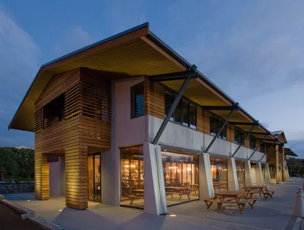 Exterior view of a contemporary two-storey timber-clad building at dusk. The building features projecting pitched roof forms, extensive ground-floor glazing, angled structural supports, horizontal timber screening and cladding, and an outdoor terrace with picnic tables. The surrounding landscape and sky are secondary to the building facade.