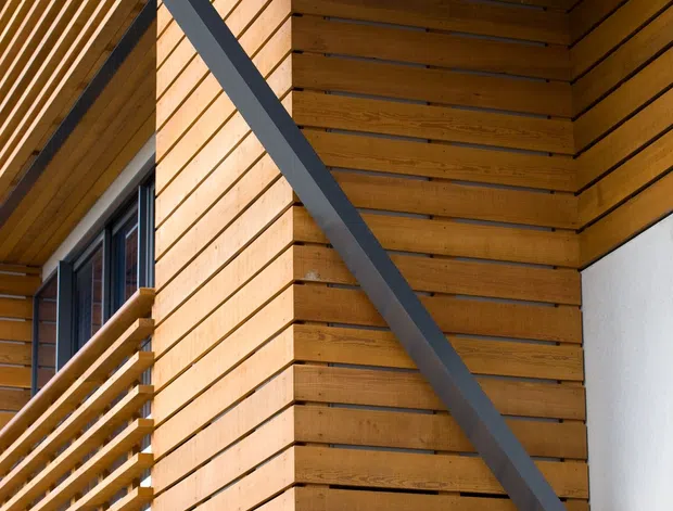 Close-up architectural exterior detail showing warm timber slat cladding and soffit panels, a timber privacy or balcony screen, and prominent dark steel diagonal and horizontal structural bracing. The composition highlights the junction of a timber-clad facade, roof overhang, glazing, and white rendered wall.