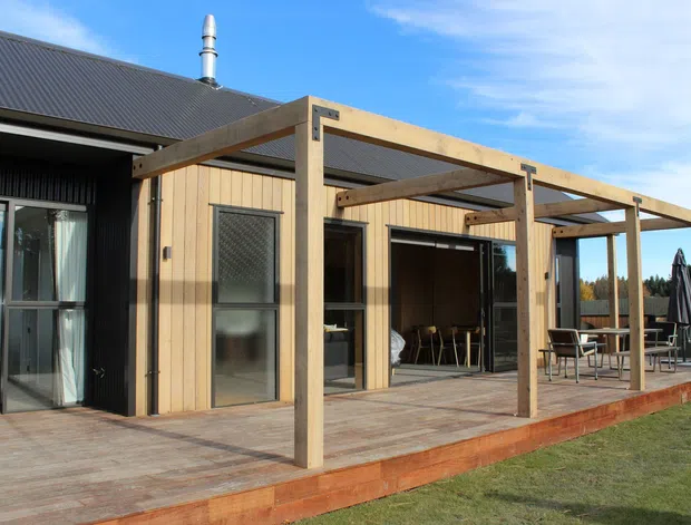 Single-storey contemporary home exterior with dark corrugated roofing and black vertical cladding combined with a warm timber-clad extension. A large timber deck and exposed timber pergola frame outdoor dining and lounge areas, with extensive sliding glazing and doors opening onto the deck.