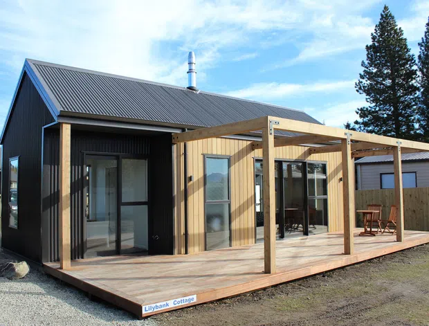 A compact single-storey cottage with dark vertical cladding, a pitched corrugated metal roof, and a light timber-clad extension opening onto a broad timber deck. The deck includes a simple timber pergola structure, with large glazed doors and windows connecting the interior to the outdoor area. The building is photographed in a residential setting with neighbouring structures and tall trees in the background.