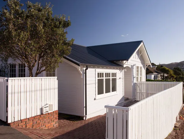 Exterior view of a compact white weatherboard house or cottage with a dark pitched gable roof, white-framed windows, brick paving, and white picket-style fencing. The building sits on a sloping suburban site with neighbouring homes and hills visible in the background.
