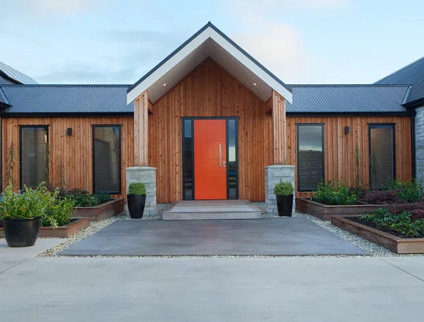 Front-facing exterior view of a contemporary single-storey building with a symmetrical gabled entrance pavilion. The facade combines vertical timber cladding, pale masonry side walls, dark metal pitched roofs, and a vivid orange-red entry door. Raised planted garden beds, potted shrubs, concrete paving, and a broad driveway frame the entrance.
