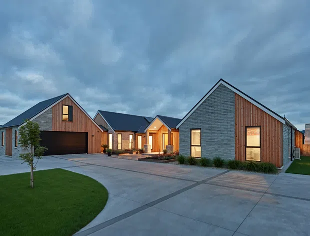 Exterior view of a contemporary low-rise residential development composed of several connected or clustered gabled volumes. The buildings combine dark grey masonry cladding, vertical warm timber cladding, steep dark metal gable roofs, and large rectangular windows. A broad concrete driveway and paths lead through landscaped lawns toward recessed illuminated entries and integrated garages under an overcast evening sky.