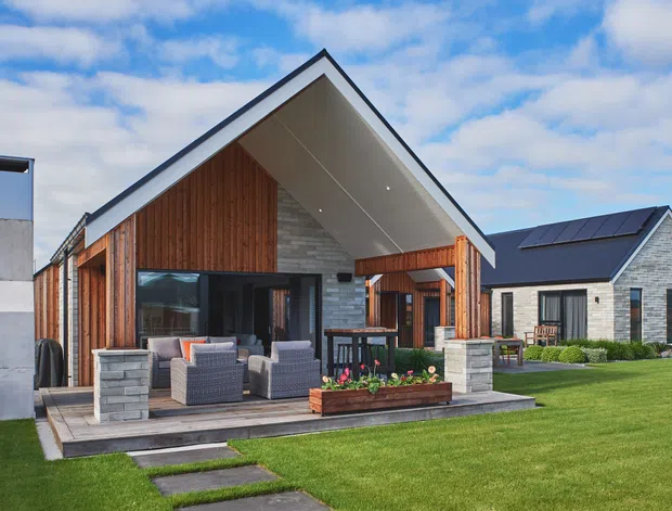 Exterior view of a contemporary single-storey home with a prominent steep gable roof, deep covered patio, timber-clad feature walls, light masonry, extensive glazing, and a landscaped lawn. Outdoor lounge furniture and a raised planter sit beneath the covered terrace; adjacent roof-mounted solar panels are visible.