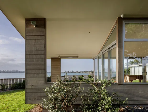 Exterior view of a low-slung modern home beside a body of water, seen from a covered terrace or verandah. The facade combines dark horizontal timber-style cladding, large aluminium-framed glazing, and a broad flat roof overhang. A glass-enclosed interior space and outdoor seating are visible, with landscaped shrubs and lawn in the foreground and a waterfront setting beyond.