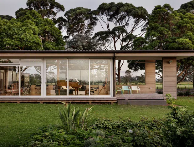 Single-storey modern pavilion-style house set within a landscaped garden, featuring extensive floor-to-ceiling glazing, a shallow flat roof, warm horizontal timber cladding, and a sheltered outdoor sitting area. The interior is visible through the glass, including a dining and living area. Mature trees and open grounds surround the building.