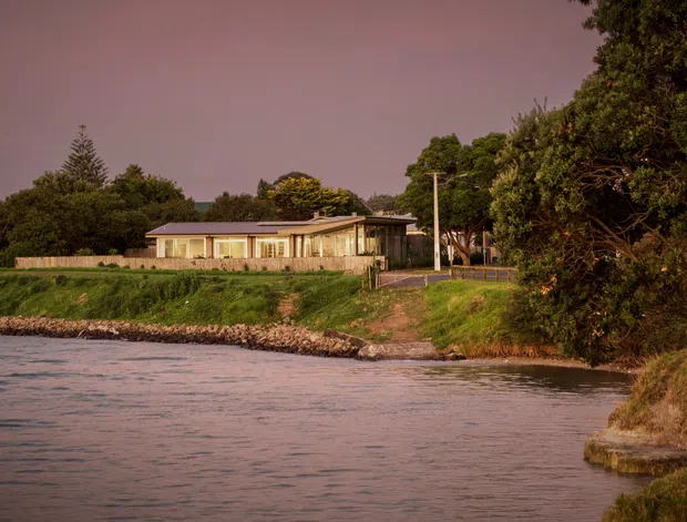 A single-storey low-profile house viewed across calm waterfront, with broad glazing, a shallow pitched roof, pale exterior walls, and a timber fence. The property sits on a gently sloping grassy bank beside the water, surrounded by mature trees and landscaping in warm dusk light.