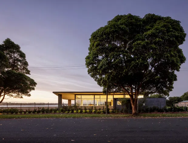 Single-storey modern residence viewed from across a quiet street at dusk. The low, horizontal building has a flat roof, extensive floor-to-ceiling glazing, warm interior lighting, and a dark masonry or rendered wing partly screened by a large mature tree. A simple landscaped frontage and linear fence complete the understated contemporary composition.
