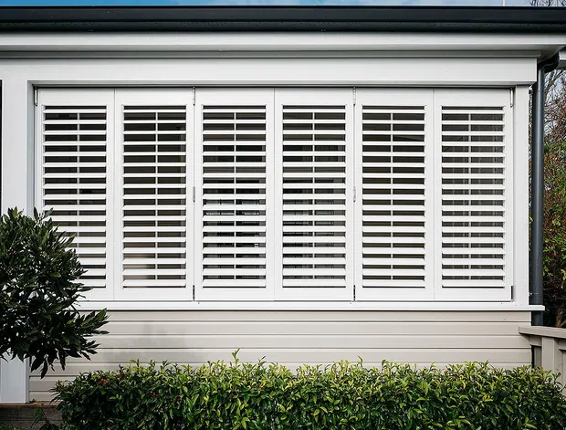 Close-up exterior view of a light-coloured weatherboard facade with a wide bank of white plantation-style louvred shutters over windows. The facade has simple horizontal trim and a dark roof edge, with hedging and shrubs in the foreground.