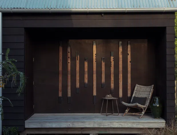 A dark timber-clad exterior facade with a recessed covered porch or entry alcove. The porch features a weathered timber deck, a large dark timber door or wall panel decorated with vertical timber elements, and a woven lounge chair with a small side table and planter. A corrugated metal skillion roof and exposed guttering complete the simple cabin-like composition, surrounded by garden planting.