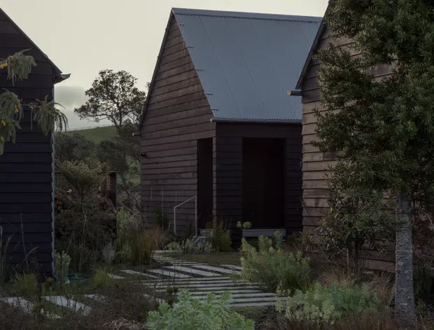 A dark, moody exterior view of a group of compact gable-roofed buildings in a landscaped setting. The buildings feature horizontal dark timber weatherboard cladding, steep pitched roofs with dark grey metal roofing, and recessed entry openings. A stepping-stone and timber walkway passes through native-style planting and shrubs between the structures. The architecture has a simple cabin-like, contemporary rural character.
