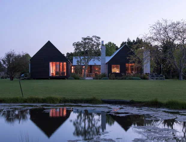 A low, dark-clad residential building viewed across a broad lawn and reflective pond at dusk. The home is composed of several steeply pitched gabled volumes with warm light glowing through large windows, connected by lower sections and surrounded by mature trees and landscaped grounds.