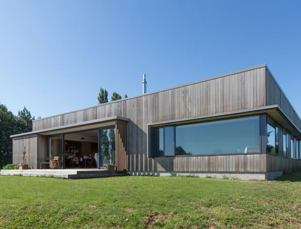 Exterior view of a low, single-storey contemporary house set in a lawned site. The building has a rectilinear flat-roof form, extensive dark-framed glazing, and vertical timber cladding across the facade. A recessed covered outdoor living area is visible on the left, while a large corner window dominates the right side.