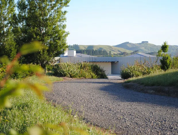 A low, contemporary single-storey building viewed from a gravel driveway, set within a landscaped rural site. The building has pale rendered walls, a corrugated metal low-pitched roof, and a large vertically ribbed or corrugated metal garage-style door. Mature trees and planting frame the foreground, with rolling hills visible beyond.