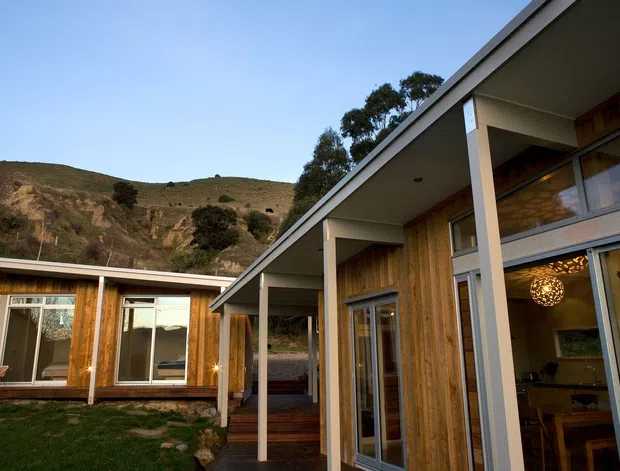 Exterior view of a contemporary single-storey home arranged around a small landscaped courtyard. The building features warm vertical timber-clad walls, extensive floor-to-ceiling glazing, a long shallow-pitched roof with deep eaves, and sheltered outdoor circulation. A rugged sloping hillside rises behind the property.
