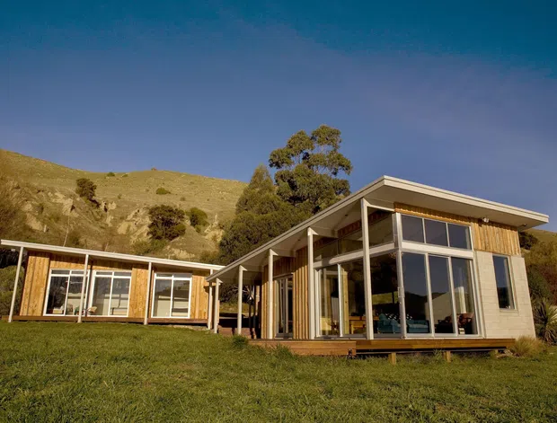 Single-storey contemporary residential house set on a grassy hillside, with a low-pitched roof, extensive floor-to-ceiling glazing, timber-clad wall sections, slender structural posts, and a projecting glazed living area. The design opens toward the landscape and has a warm timber-and-glass character.