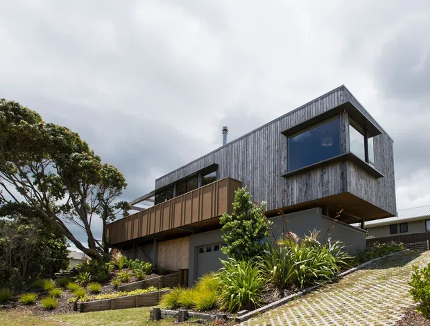 Exterior view of a contemporary multi-level house on a sloped site, featuring dark weathered timber cladding, projecting box-like upper volumes, large windows, a broad timber balcony, and an integrated garage. Landscaped planting and a steep paved driveway occupy the foreground beneath an overcast sky.