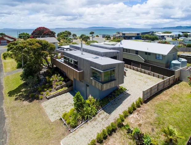 Aerial view of a contemporary coastal residential property, featuring a compact two-storey home with dark vertical timber-style cladding, a flat roof, extensive paved outdoor areas, and landscaped gardens. The surrounding suburban neighbourhood and broad waterfront setting are clearly visible.
