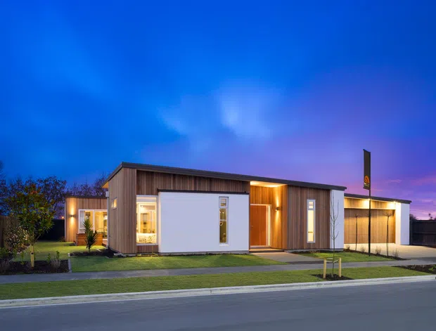 A single-storey contemporary residence photographed at dusk from the street. The compact home has a low-pitched flat roof form, warm vertical timber cladding, white rendered wall panels, extensive glazing, a recessed illuminated entry, and an attached garage. A neat lawn, paved path, driveway, and young landscaping sit in front of the house.