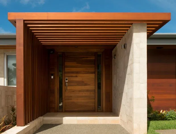 A contemporary residential entrance viewed from outside, featuring a broad timber pergola with closely spaced slatted rafters, a timber-clad front door and sidelights, a pale stone-clad structural wall, and a textured concrete pathway and steps. The entry is framed by lawn and planting beneath a bright blue sky.