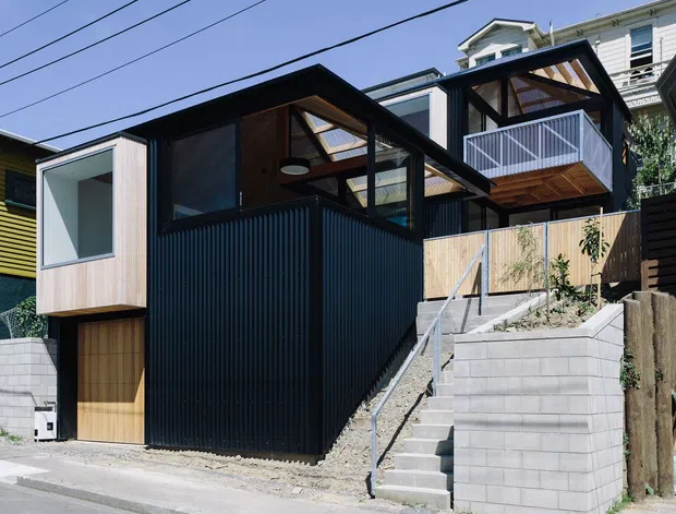 Street-level exterior view of a compact contemporary multi-level dwelling on a sloped urban site. The house features dark vertically ribbed cladding, timber soffits and balcony decking, large glazed openings, a timber garage door, and an external concrete stair leading to a raised entrance and balcony. Overhead power lines and neighbouring homes are visible.