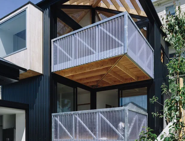 Exterior view of a contemporary multi-storey house with a prominent open timber gable roof structure, black vertical cladding, pale timber wall cladding, and two stacked balconies enclosed by translucent corrugated screens. The lower level includes glazed openings, a wood storage area beneath the balcony, patterned permeable paving, and a sloped landscaped side path.