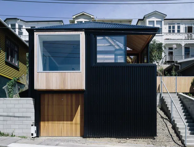 Street-facing view of a compact contemporary residential extension or studio, clad in dark vertical metal siding with a timber garage door and light timber-framed projecting window. The building has a low-pitched corrugated roof, a partially open upper-level terrace or covered outdoor space, and concrete steps rising along the right side. Older multi-storey homes are visible behind it on a dense sloping suburban street.