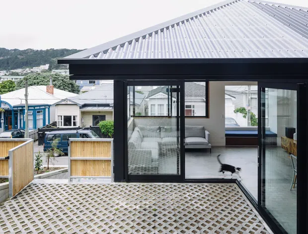 Exterior view of a contemporary dark-clad home extension with a large light-grey corrugated metal pitched roof and extensive black-framed glazing. The elevated paved terrace has a patterned concrete grid surface and timber privacy fencing. Through the glazing, a bright living area with a sofa, armchair and low furniture is visible. A suburban neighbourhood and wooded hillside appear in the background.