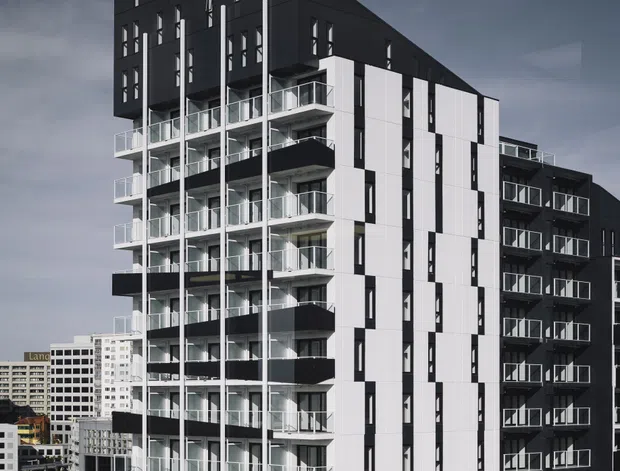 A tall contemporary multi-storey apartment building in an urban setting, photographed from outside. The facade combines dark charcoal cladding with white vertical panels, extensive glazed balconies, narrow vertical windows, and a sharply angled roof form. Surrounding city buildings are visible in the background under an overcast sky.