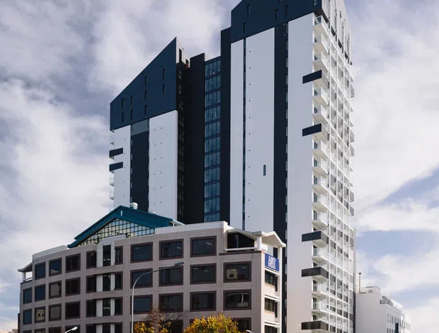 Street-level exterior photograph of a contemporary multi-storey urban apartment or mixed-use development. Two tall, sharply angular tower volumes with dark cladding and white vertical facade panels rise behind a lower masonry-clad building with a distinctive glazed pitched roof. Repetitive balconies line one tower, while surrounding streets, traffic lights, pedestrians, and mature street trees establish a dense city setting.