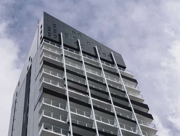 Low-angle exterior photograph of a tall contemporary multi-storey apartment building. The facade combines dark grey metal or panel cladding with repetitive projecting balconies, transparent glass balustrades, vertical structural or facade elements, and small square windows. The image is dominated by the building against a cloudy sky.