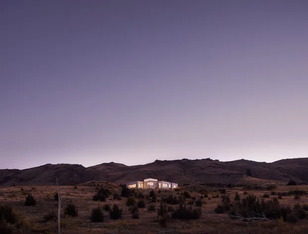 A distant view of a compact illuminated house set in a rugged, sparsely vegetated landscape at dusk, with dark rolling hills behind it and a broad purple-grey sky. The building appears contemporary and low-rise, but facade details are limited by distance and low light.