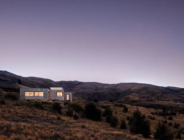 A small, contemporary single-storey house set in a rugged, sparsely vegetated hillside landscape at dusk. The compact rectangular building has dark grey exterior walls, large warm-lit windows, and a simple low-pitched or flat roof. Rolling brown hills dominate the background, creating a remote rural setting.