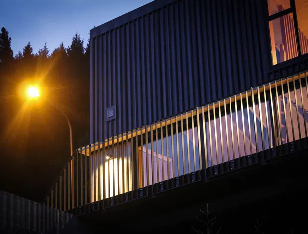 A dusk exterior view of a contemporary multi-level building, featuring dark vertically ribbed cladding, illuminated glazing, and a projecting balcony or terrace enclosed by slender vertical metal balustrades. Warm interior lighting contrasts with the cool blue evening light and surrounding trees.