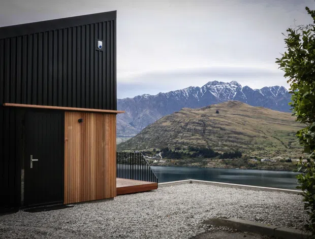 Exterior view of a compact modern dark-clad structure with vertical ribbed black wall panels, a timber slatted entry enclosure, and a small timber deck beside a gravel forecourt. A lake and dramatic snow-dusted mountain range are visible beyond the property, creating a strong alpine setting.