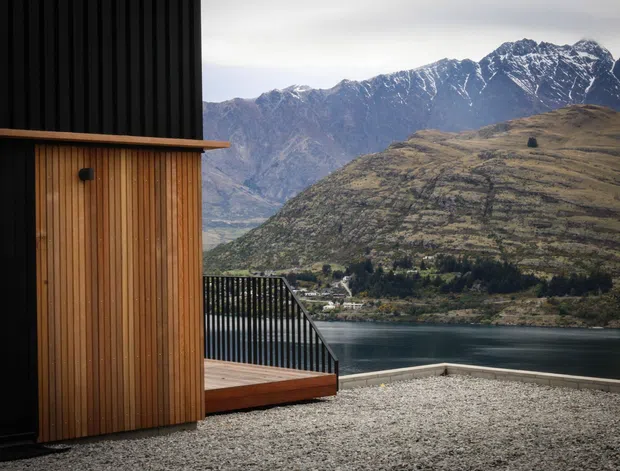 Exterior architectural view of a modern dark-clad building with a warm vertical timber-clad wall and timber deck, overlooking a lake and rugged snow-capped mountains. A black metal balustrade and gravel foreground reinforce the contemporary alpine setting.